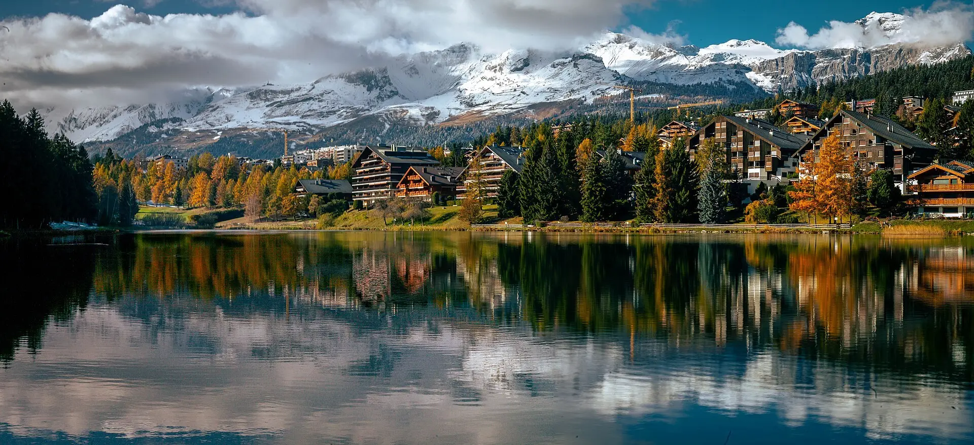 A couple on a lakeside path in Crans-Montana, pine trees around them and the snow peaks of the Valais Alps across the Rhône valley