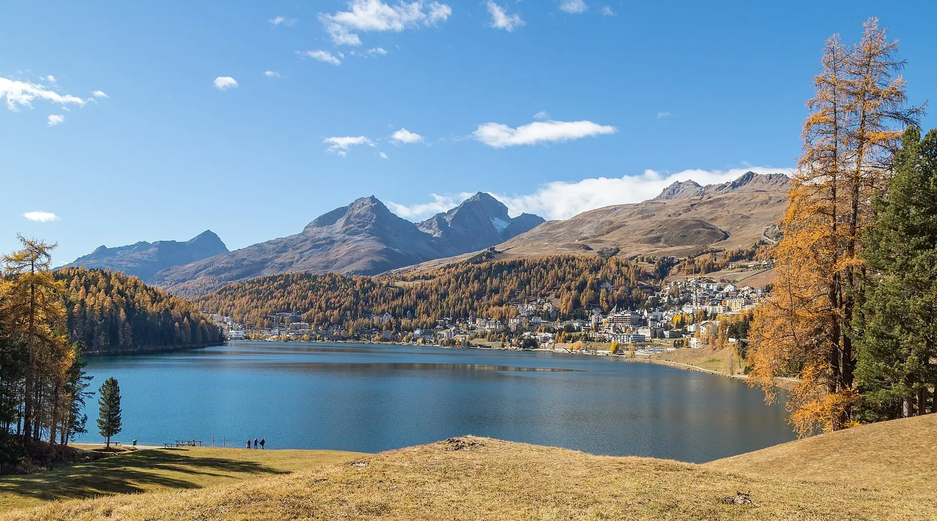 A honeymoon couple on a hotel terrace above St. Moritz in late afternoon light, snow-covered peaks behind them